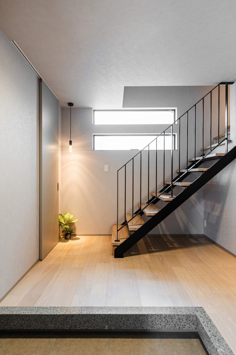 石目調の上がり框とアイアンの質感が、外観に採用した大谷石調タイルや随所に施された細部のデザインと美しく呼応する玄関ホール。内と外で素材のトーンを揃えることで、住まい全体が統一された印象になります。<br>明るさを確保するために高窓とストリップ階段で、抜け感と自然な明るさを演出。グレイッシュなトーンでまとめ、デザイン性のあるペンダントライトが映えるシンプルな空間です。