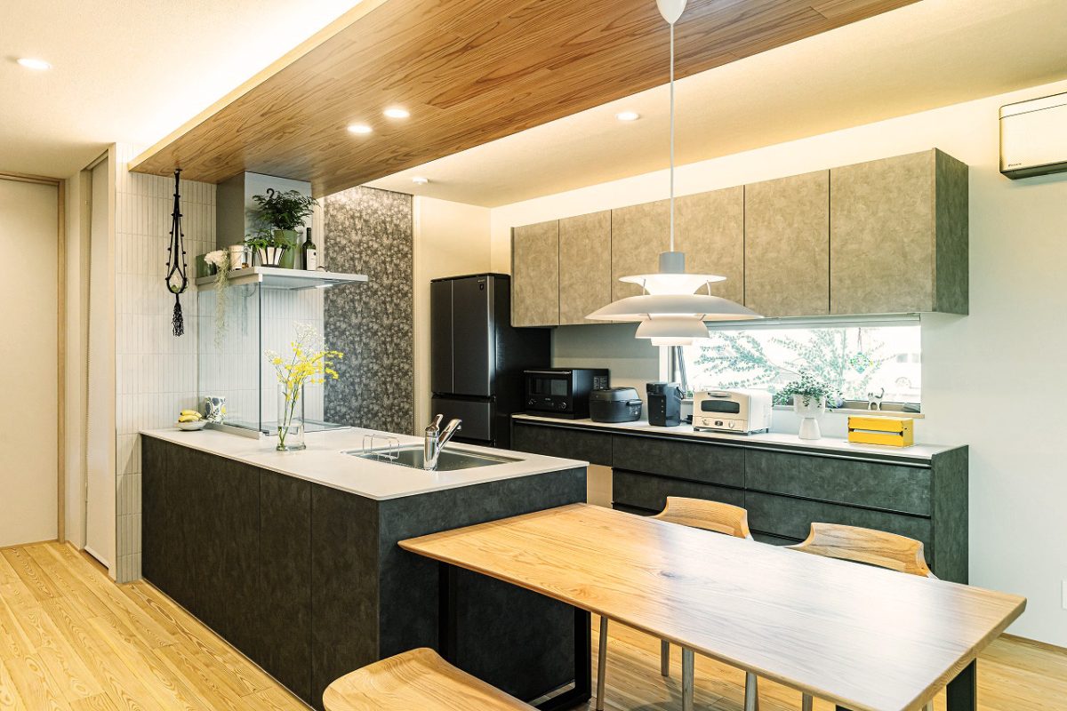 カップボードの上部はトーンアップしたグレーにすることで、暗くなりすぎないようにコーディネートしました。<br>上部には下がり天井を設け、間接照明を仕込むことで落ち着いた雰囲気を演出しています。ペンダントライトが食卓により一層彩りを添えます。<br>奥にはロールスクリーンで目隠しした、十分な収納量のあるパントリーを設けました。