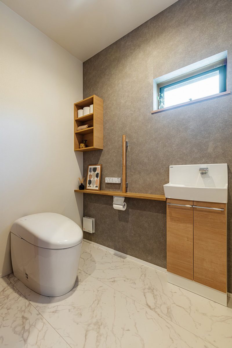 大理石調のフロアに、グレージュのアクセントクロスを合わせた上質なトイレ空間。<br>収納棚やカウンターは木目の面材を使用し、木の温もりを感じる印象にまとめました。高窓はプライバシーに配慮しつつ、自然光を取り入れることができます。