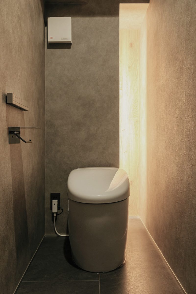 間接照明の柔らかな光に包まれた、上品で落ち着きのあるトイレ。<br>奥の壁にはスリットがあり、トイレットペーパーなどをしまえる収納スペースになっています。壁と一体化させることで、デザイン性を損なうことなく、洗練された仕上がりにしました。