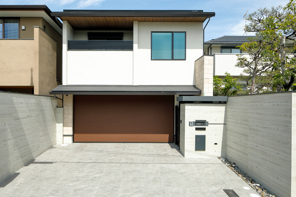 外壁はベージュの吹付けとタイル、ウォルナット木目調の軒天と電動シャッターで重厚感を演出。<br>コンクリ―ト転写の塀がより印象を引き立てています。